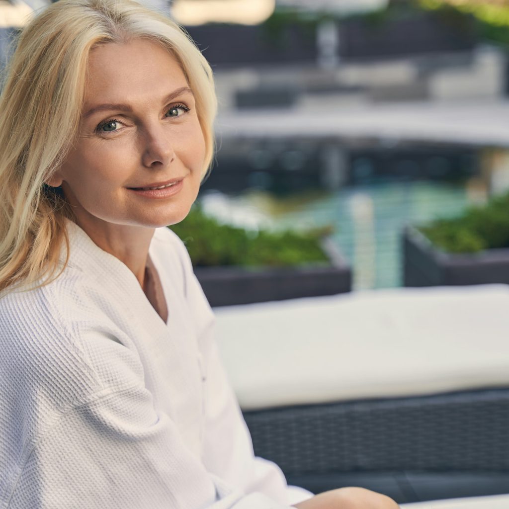 Blonde woman in her fifties in a white robe on a sunlit terrace