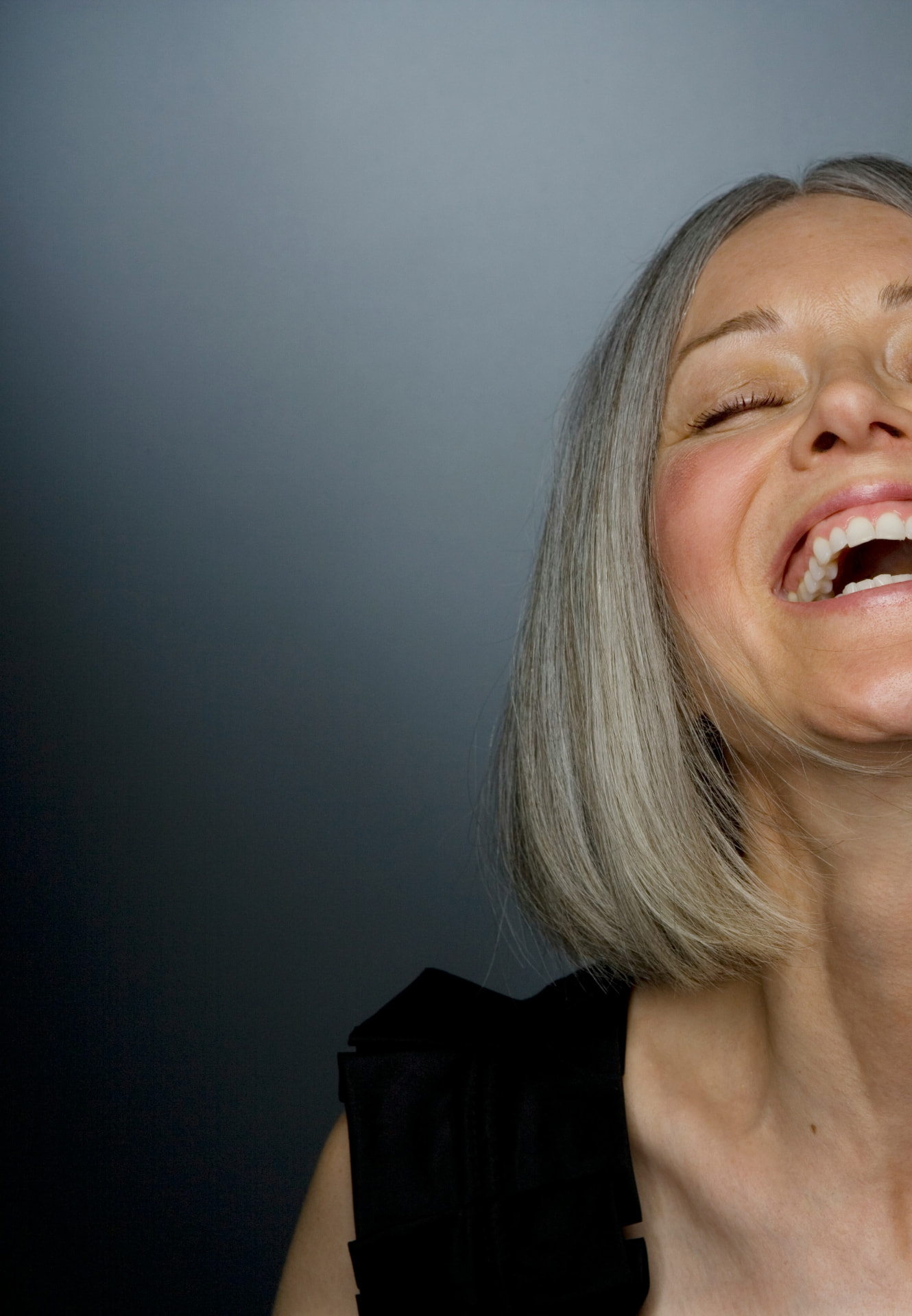 A woman with grey hair laughing, head tilted back, against a deep grey backdrop